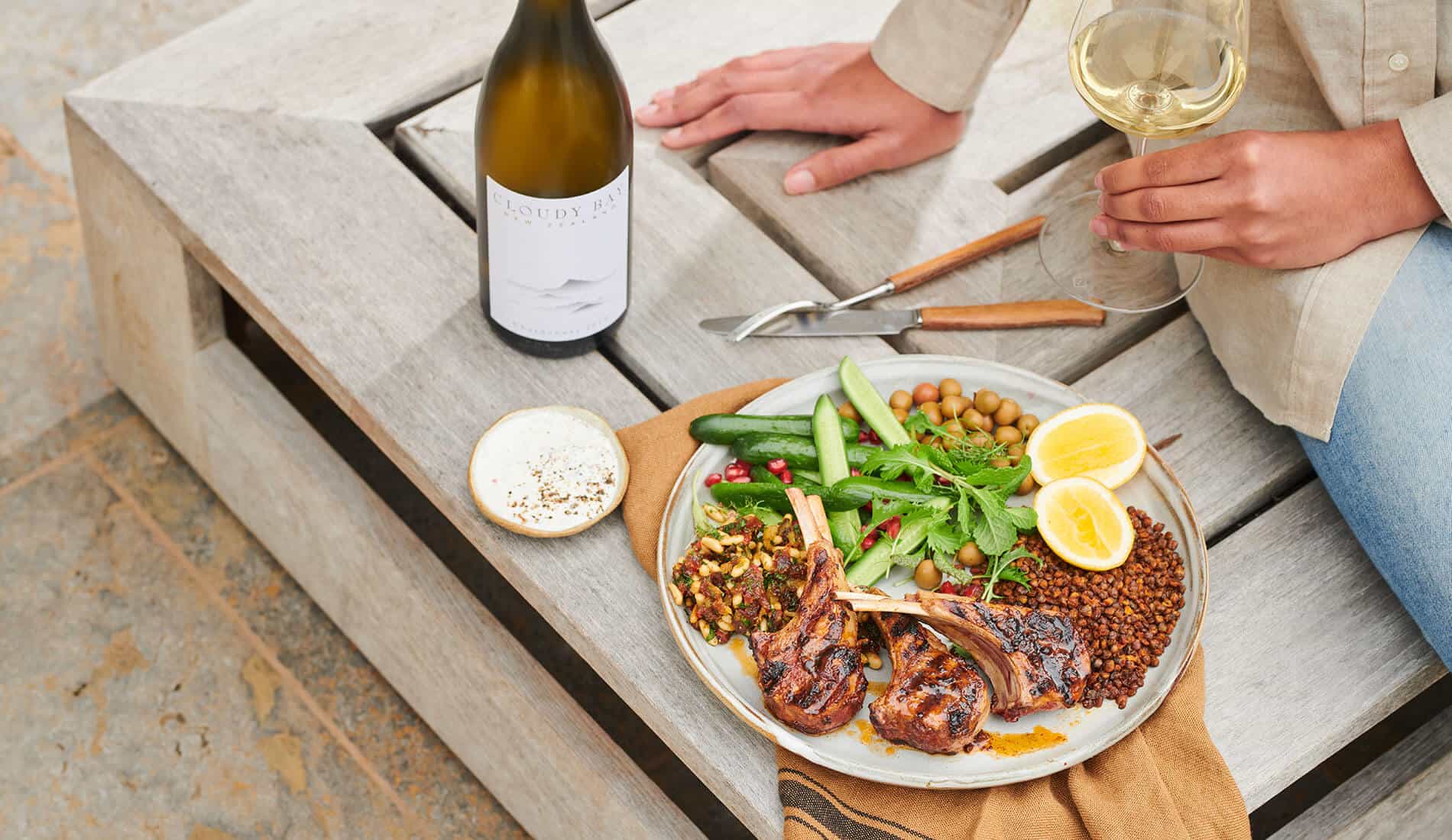 Hand holding glass of white wine beside plate of food and Cloudy Bay Chardonnay bottle