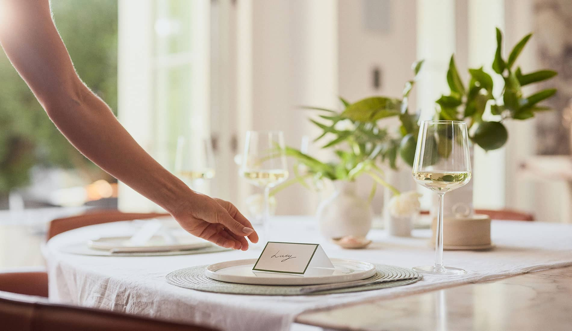 An arm in the process of setting down a name card at a table set with a glass of white wine and green foliage. The name card says Lucy.