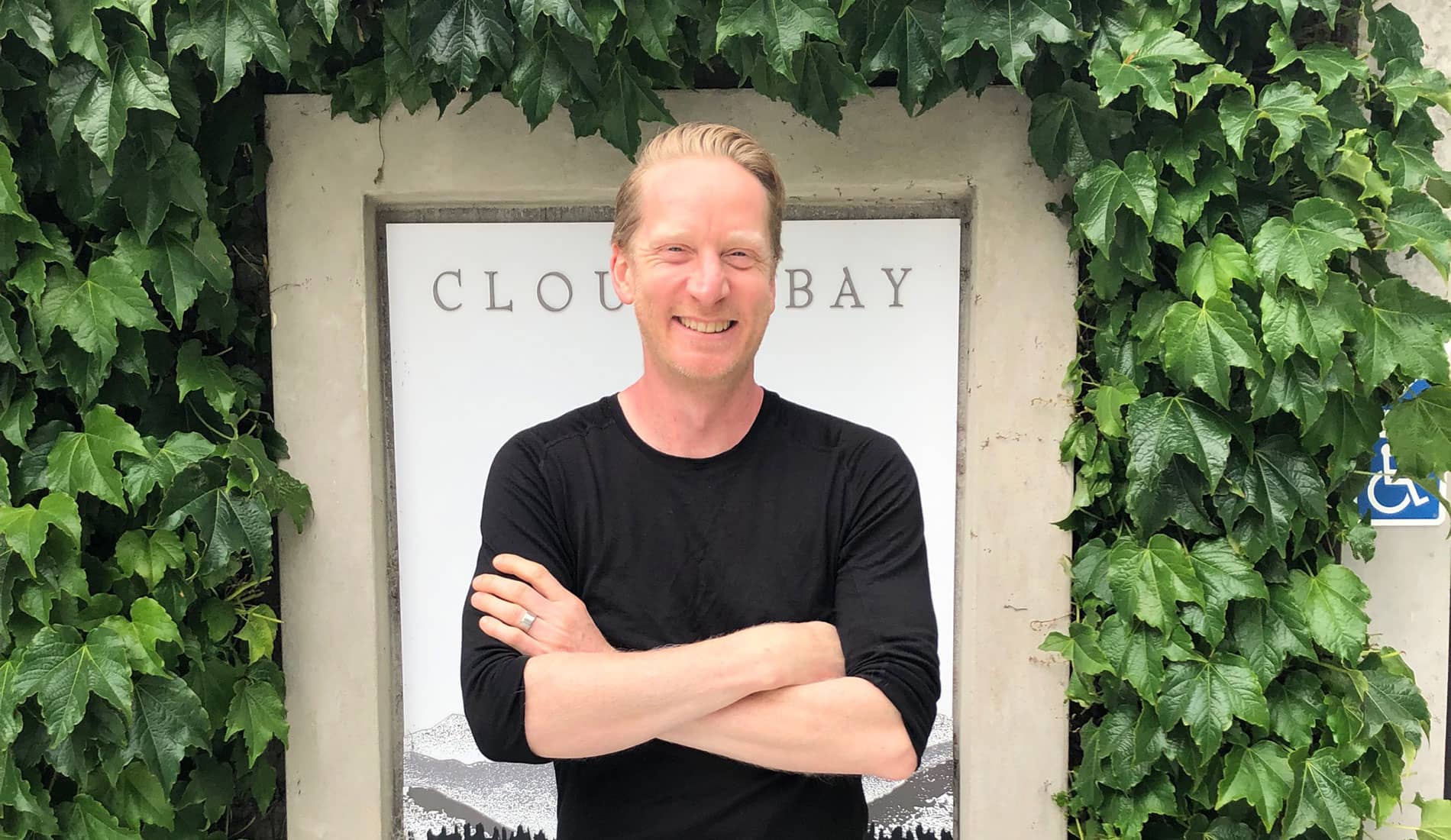 A man stands in front of the Cloudy Bay sign. His arms are folded and he is smiling.