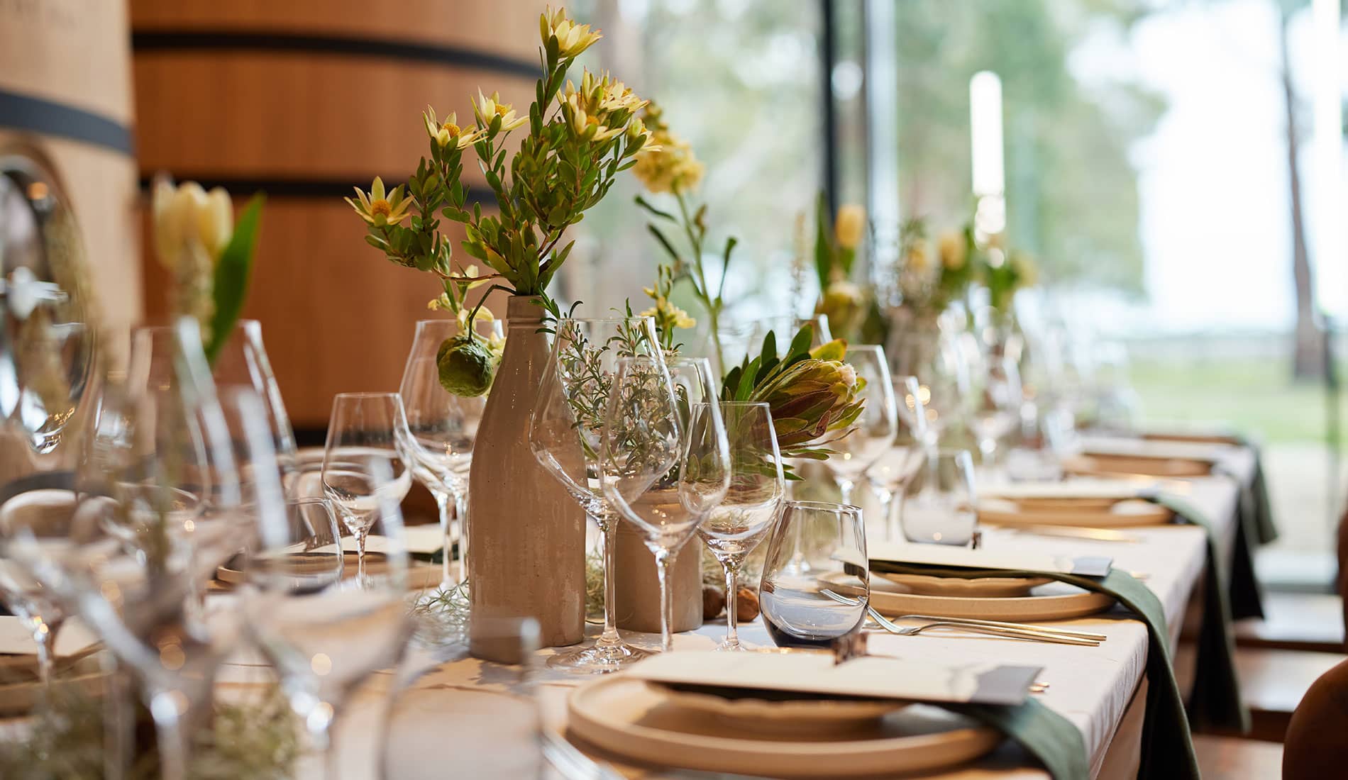 A long table set with flowers in vases and wooden wine vats in the background