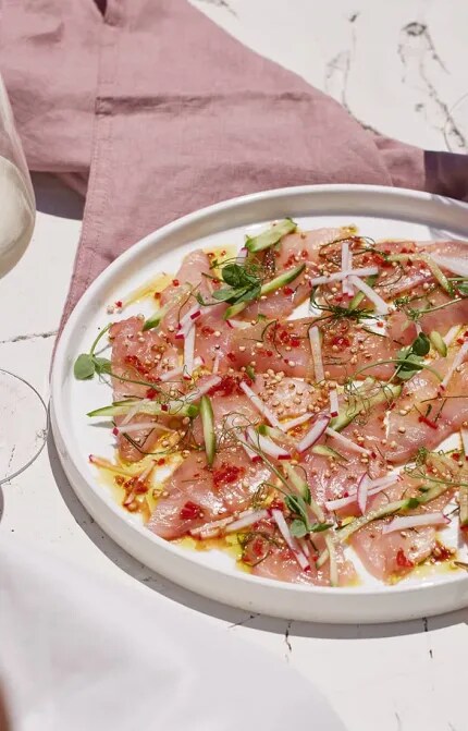 Person holding glass of white wine beside plate of raw fish and bottle of Cloudy Bay Sauvignon Blanc