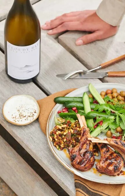 Hand holding glass of white wine beside plate of food and Cloudy Bay Chardonnay bottle