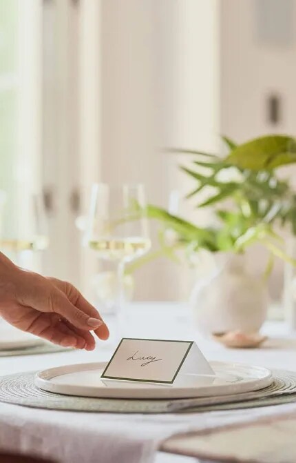 An arm in the process of setting down a name card at a table set with a glass of white wine and green foliage. The name card says Lucy.